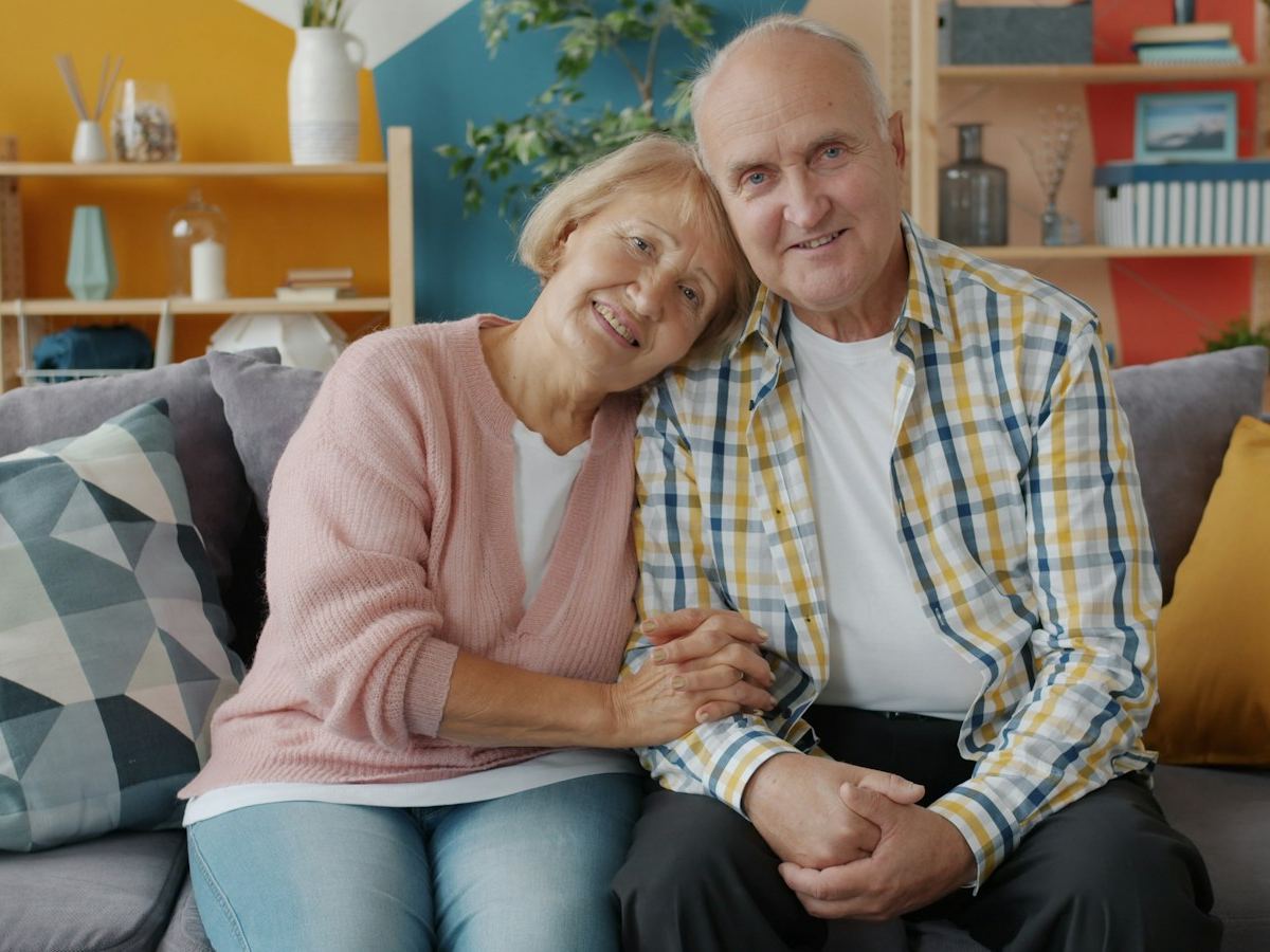 Älteres Ehepaar sitzt lächelnd und eng beieinander auf dem Sofa im eigenen Wohnzimmer